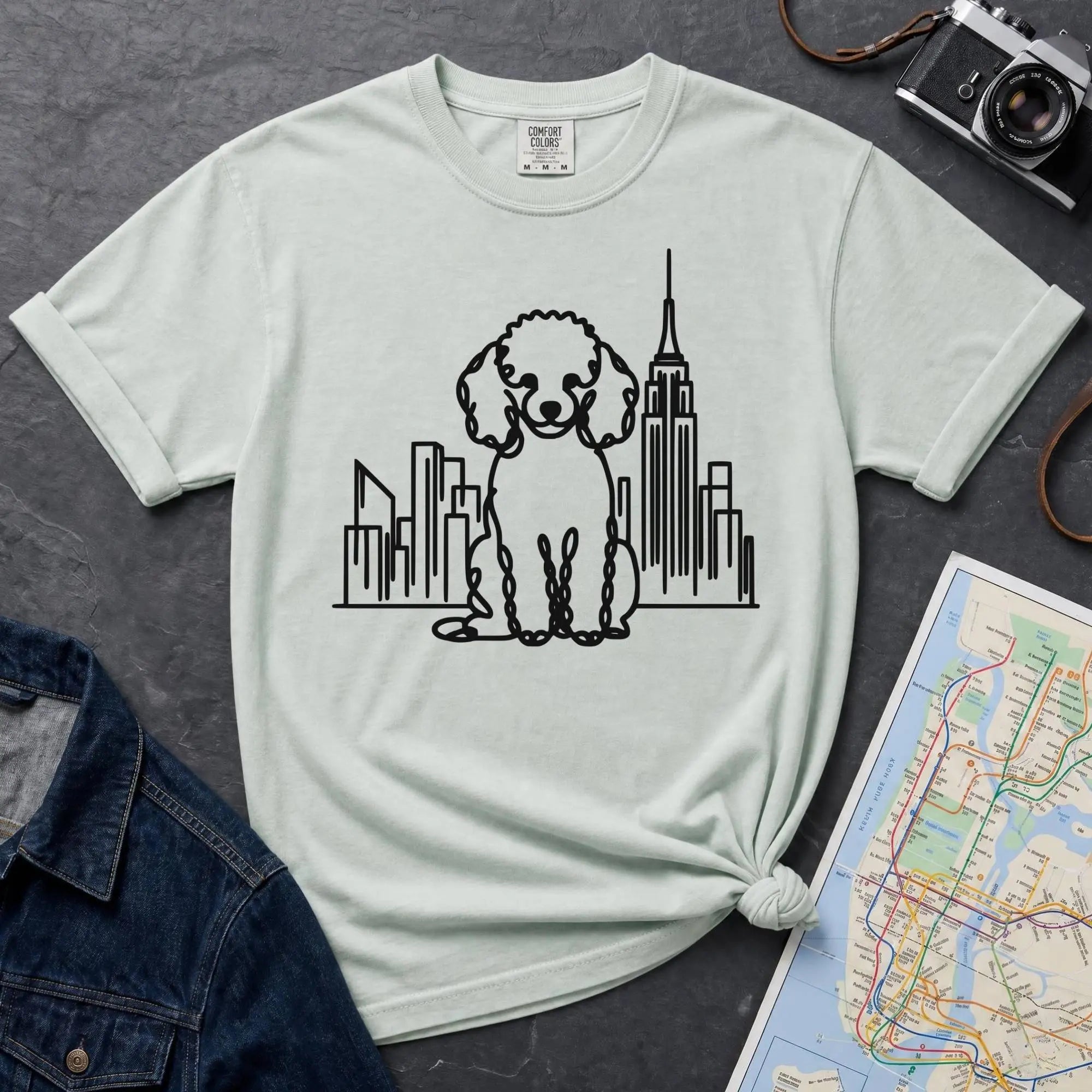 Bay colored t-shirt with a poodle and cityscape design on a dark surface with a map and camera.