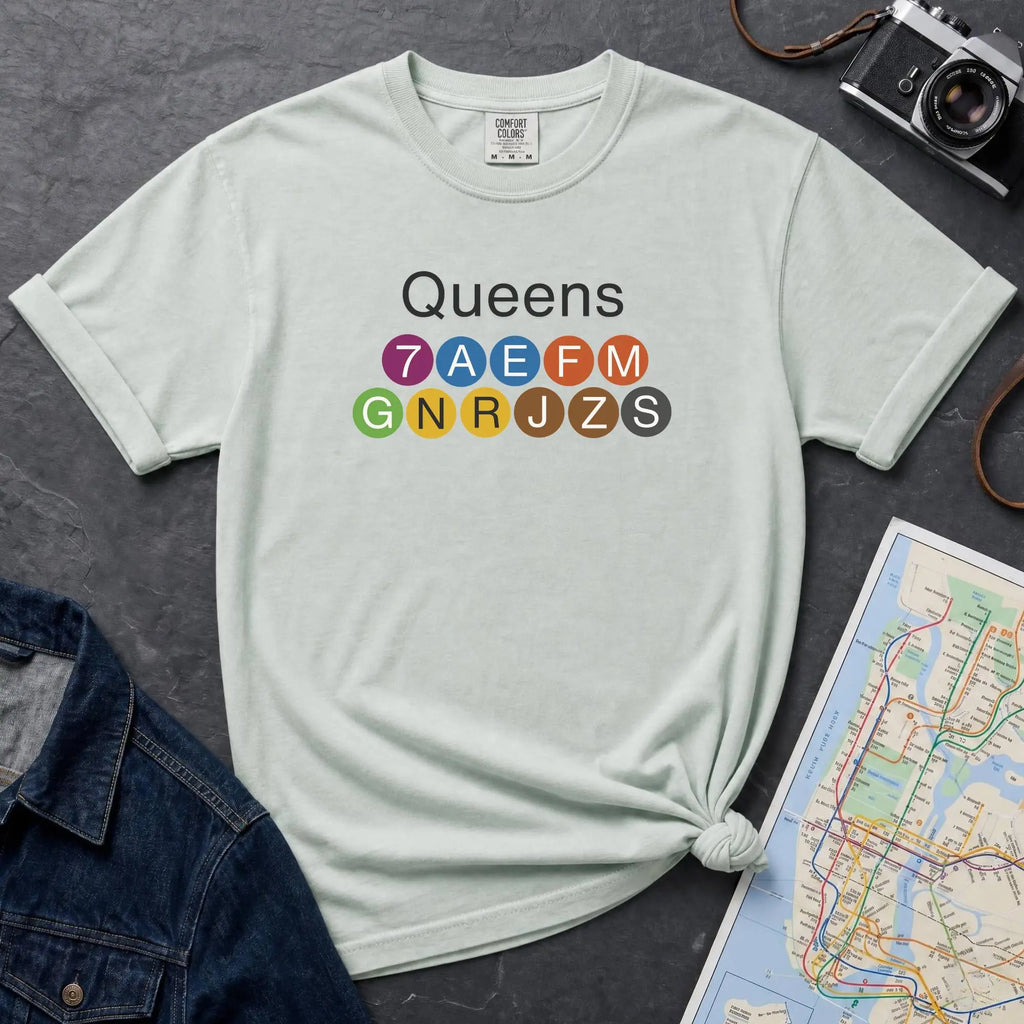 Bay colored t-shirt with 'Queens' and NYC subway lines on a dark surface with a map and denim jacket.