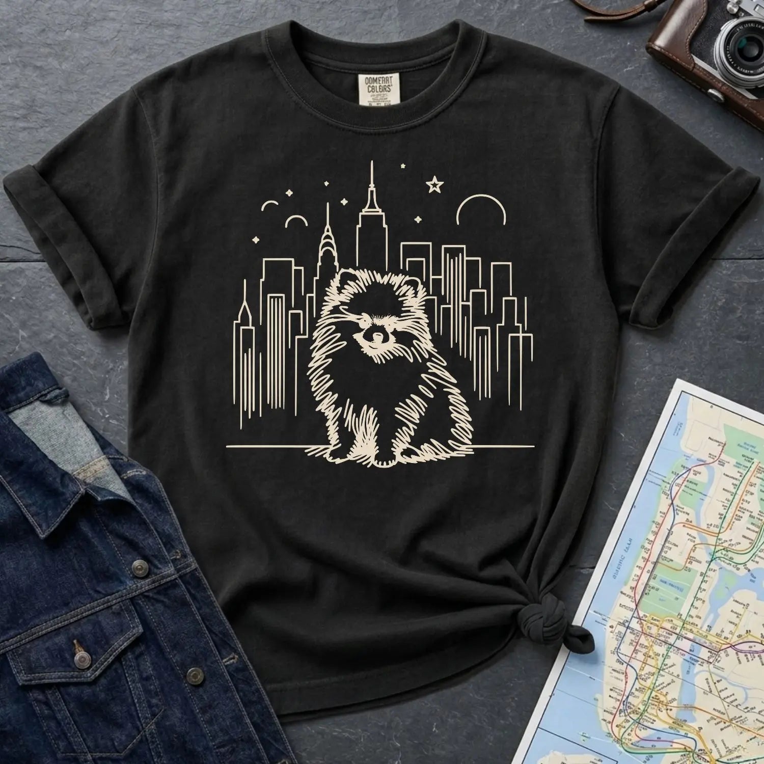 Black Comfort Colors t-shirt featuring a fluffy Pomeranian dog illustration in front of the New York City skyline on a dark surface with denim jacket and map.