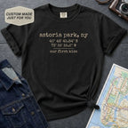Black Comfort Colors t-shirt with text 'astoria park, ny', coordinates and 'our first kiss', placed on a dark surface with a map and denim jacket.