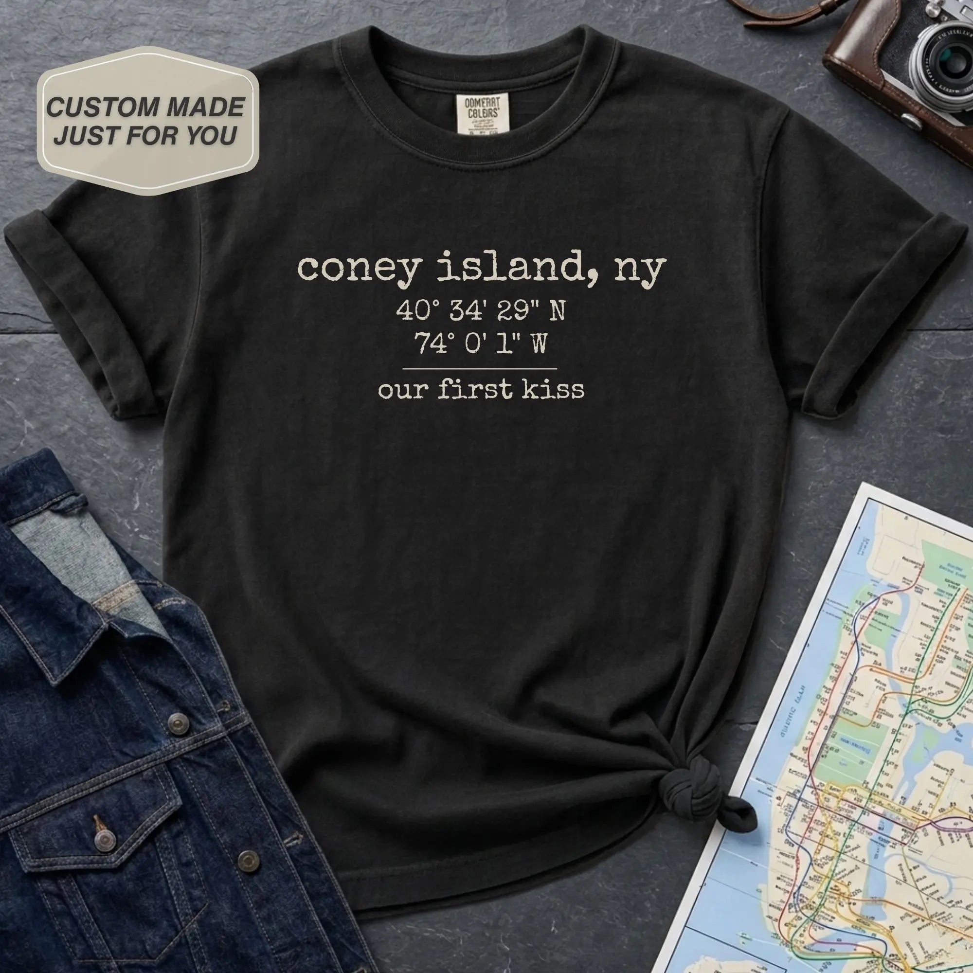 Black t-shirt with text 'coney island, ny 40°54'29"N 74°0'1"W our first kiss' on a dark surface with a map and denim jacket.