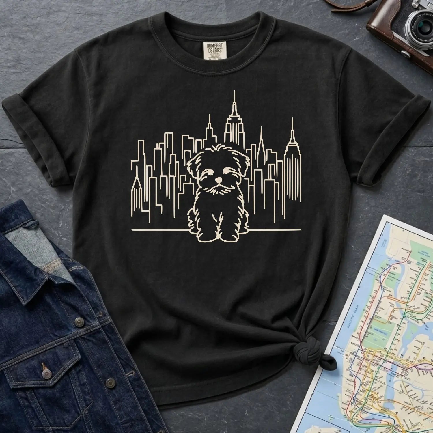 Black Comfort Colors t-shirt with a Maltese dog and cityscape design on a gray surface with a map and camera.
