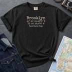 Black Comfort Colors t-shirt with 'Brooklyn' and coordinates printed on it, placed on a dark surface with a map and denim jacket.