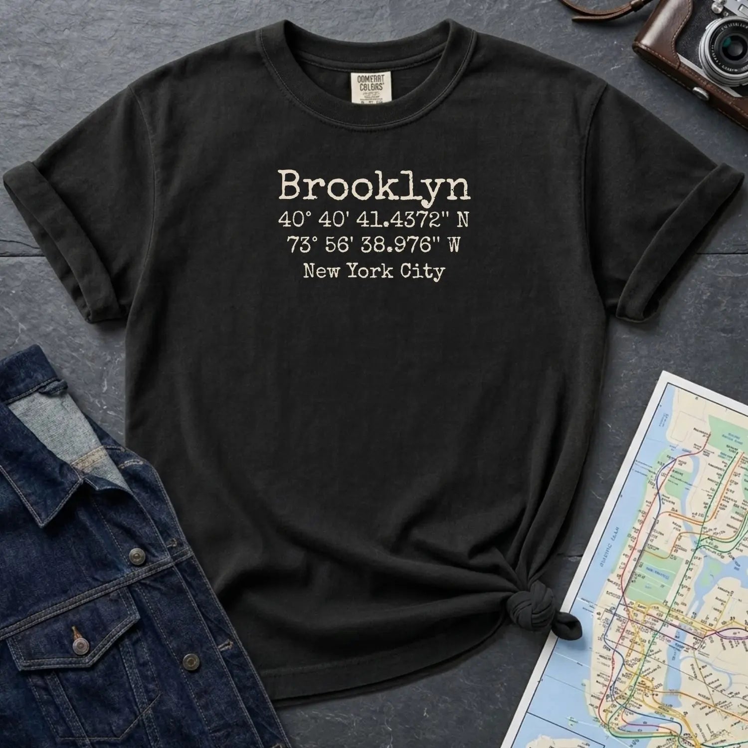 Black Comfort Colors t-shirt with 'Brooklyn' and coordinates printed on it, placed on a dark surface with a map and denim jacket.