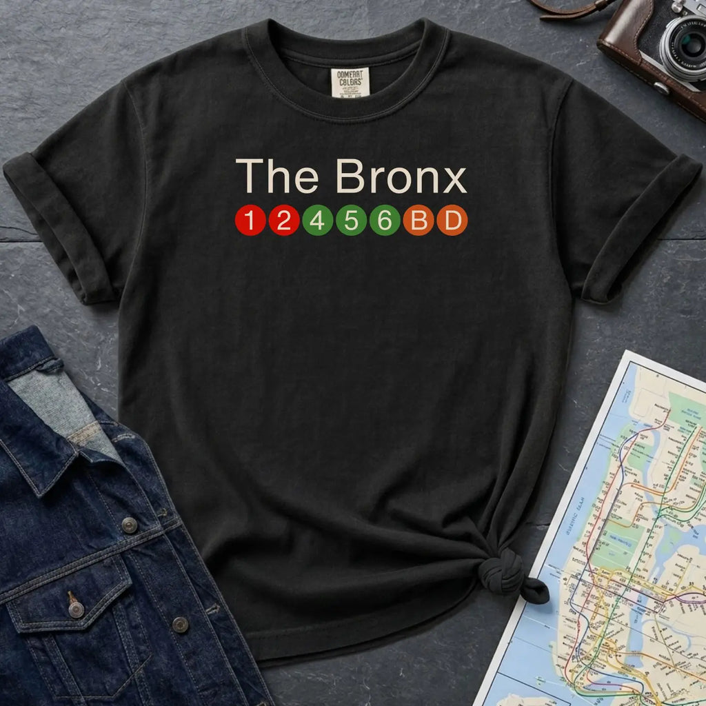 Black Comfort Colors t-shirt with 'The Bronx' text and subway line design on a gray surface with a map and denim jacket.