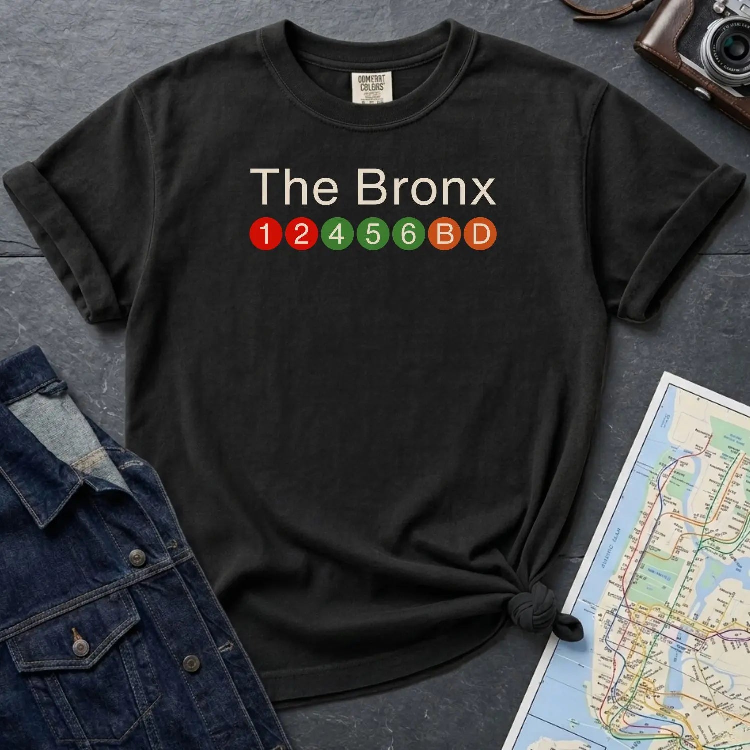Black Comfort Colors t-shirt with 'The Bronx' text and subway line design on a gray surface with a map and denim jacket.