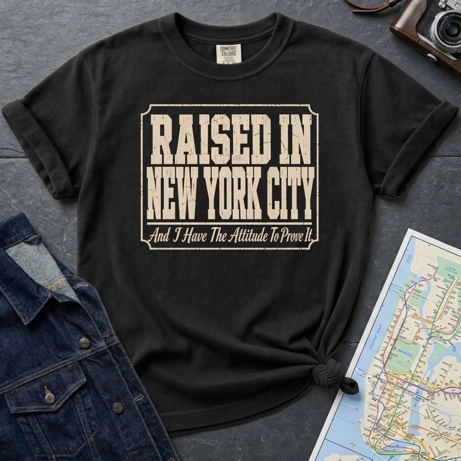Black Comfort Colors t-shirt with 'Raised in New York City' text on a dark surface with a map and denim jacket.