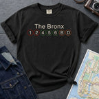 Black Comfort Colors t-shirt with 'The Bronx' and subway line text on a dark surface with a map and jacket.