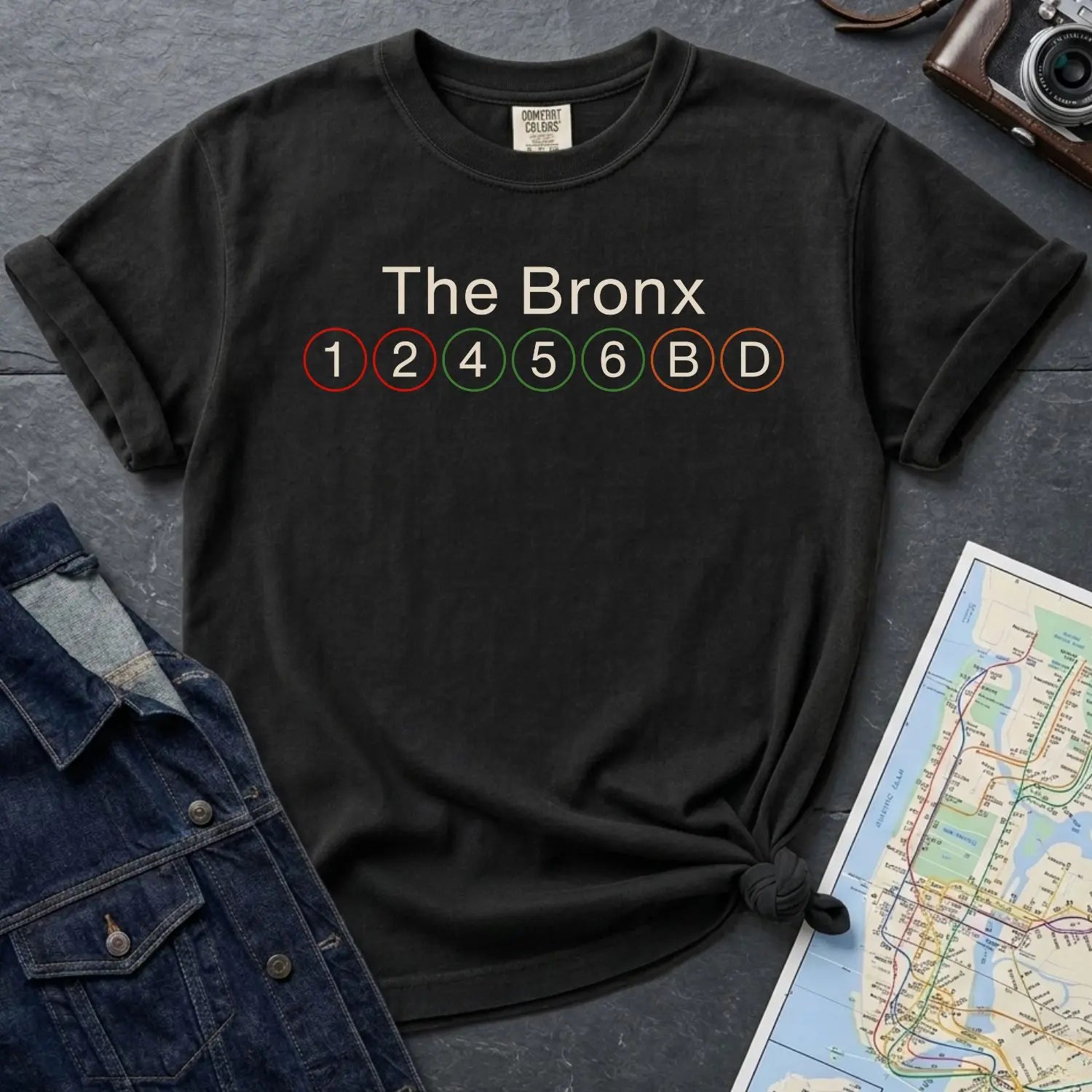 Black Comfort Colors t-shirt with 'The Bronx' and subway line text on a dark surface with a map and jacket.