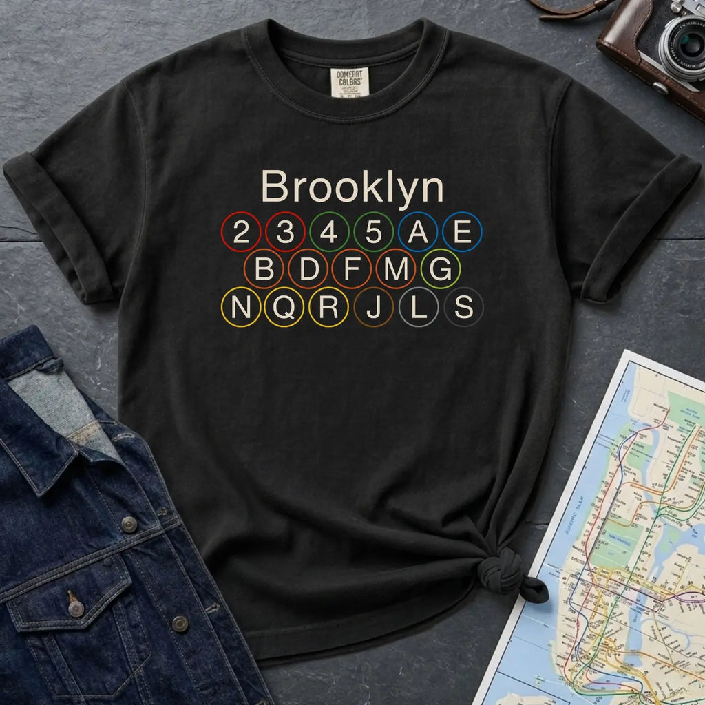 Black Comfort Colors t-shirt with Brooklyn subway line design, next to a denim jacket and map on a dark surface