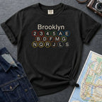 Black Comfort Colors t-shirt with Brooklyn subway line design, next to a denim jacket and map on a dark surface