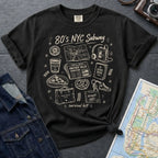 Black Comfort Colors t-shirt with '80s NYC Subway Survival Kit' graphic design on a dark surface with denim jacket and map.