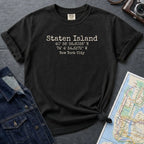 Black Comfort Colors t-shirt with Staten Island text and coordinates on a dark surface with denim jacket and map.