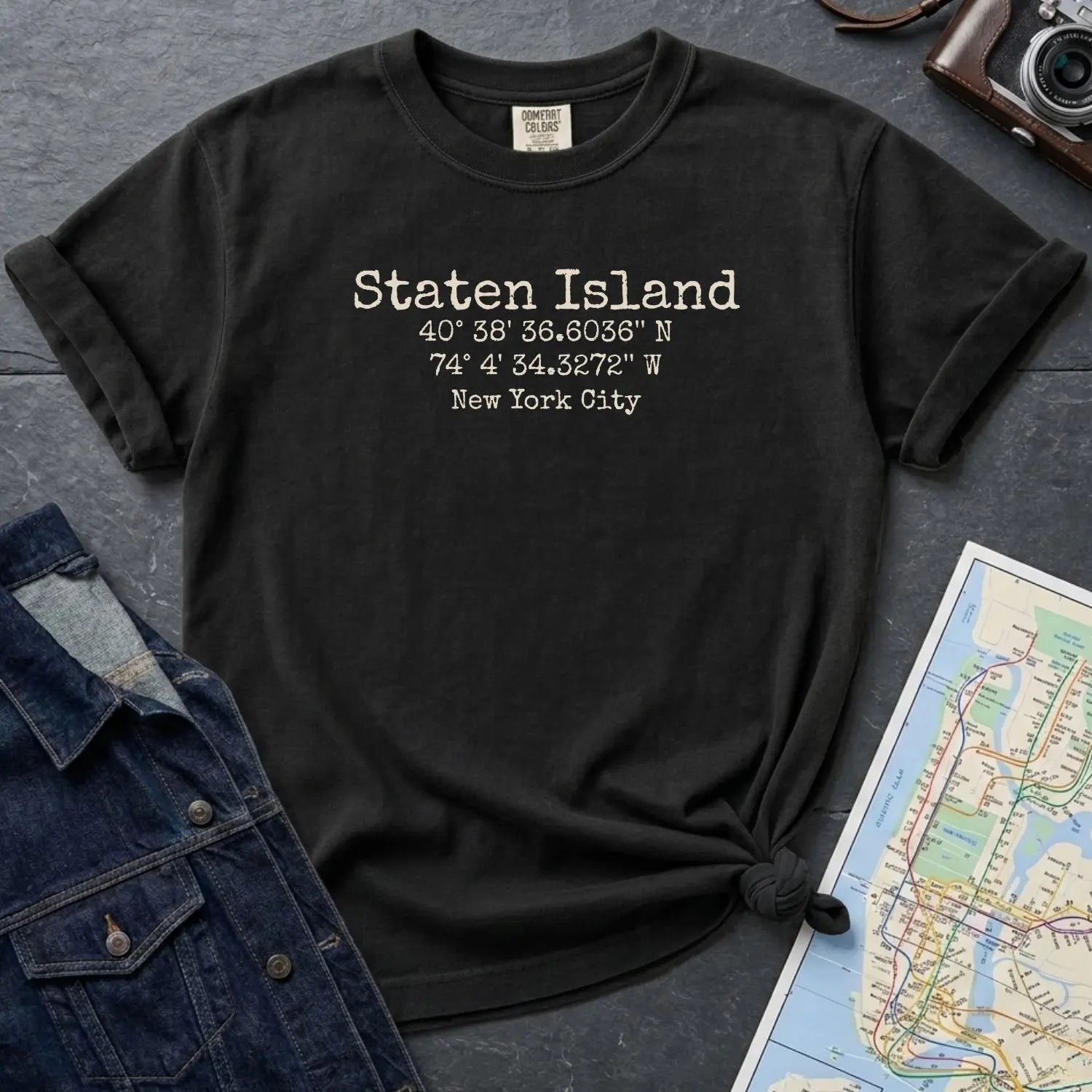 Black Comfort Colors t-shirt with Staten Island text and coordinates on a dark surface with denim jacket and map.