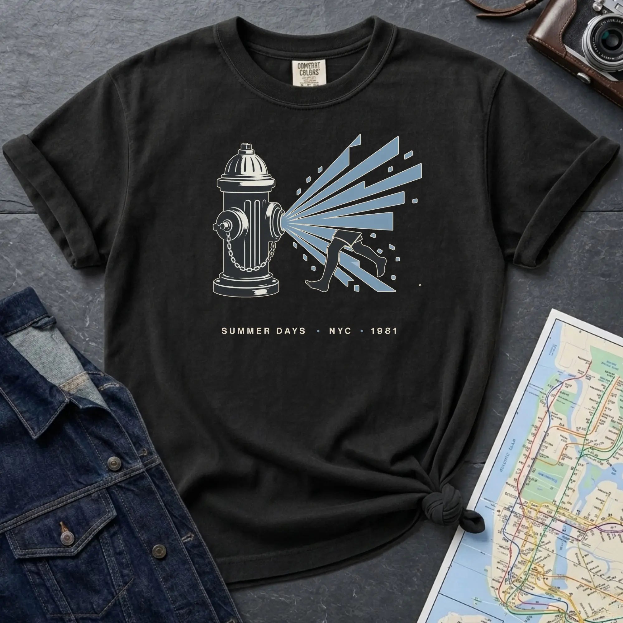 Black Comfort Colors t-shirt featuring a vintage NYC fire hydrant spraying water with “Summer Days • NYC • 1981” text, placed on a dark surface with a denim jacket and a map.