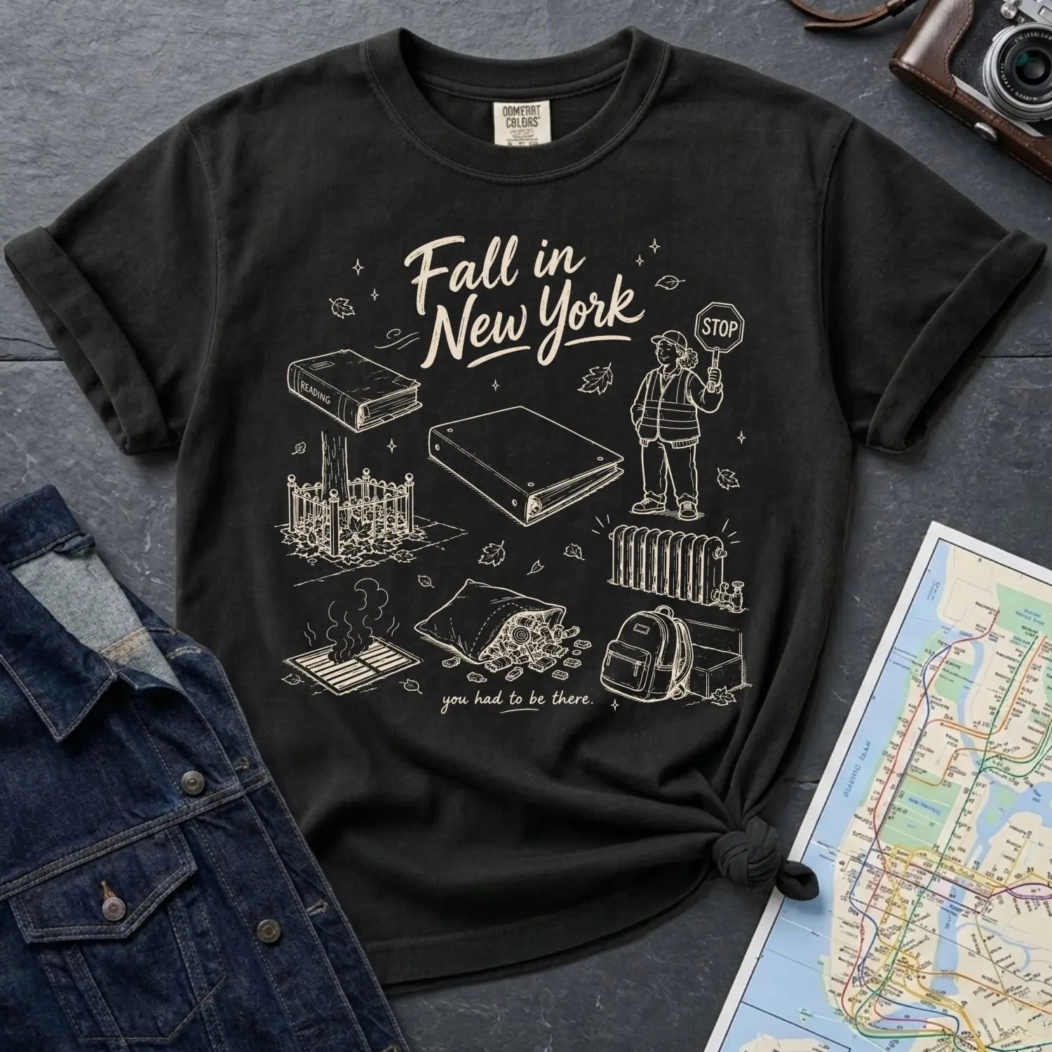 Black Comfort Colors t-shirt with 'Fall in New York' graphic design on a dark surface with a map and denim jacket.