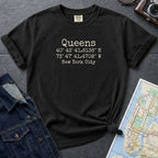 Black Comfort Colors t-shirt with 'Queens' text and coordinates, placed on a dark surface with a map and denim jacket.