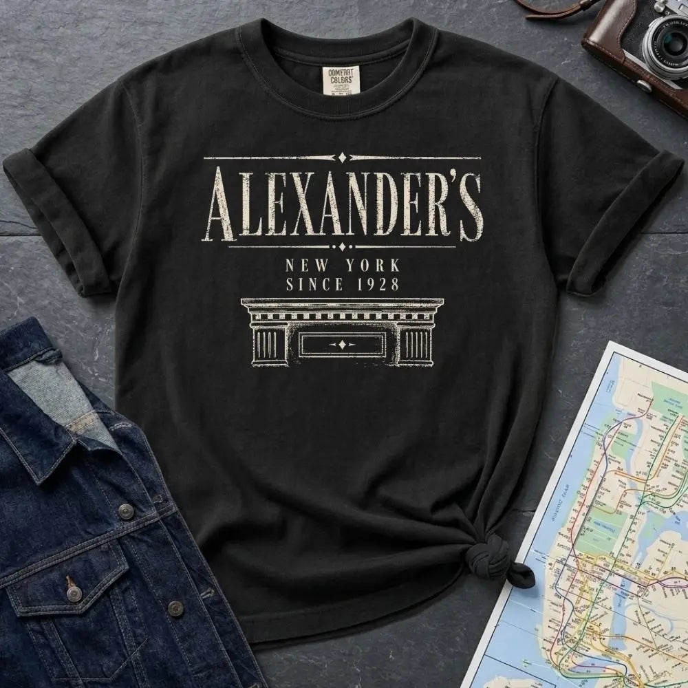 Black Comfort Colors t-shirt with vintage serif lettering reading Alexander's, New York since 1928, above an illustrated stone building cornice, styled after a classic 1920s Bronx department store facade.