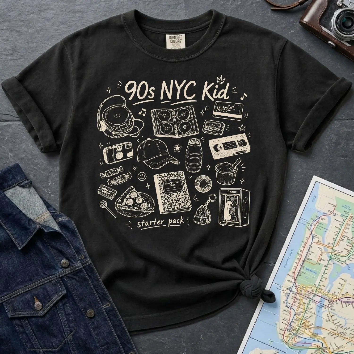 Black Comfort Colors t-shirt with '90s NYC Kid' graphic design on a dark surface with denim jacket and map.