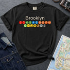 Black Comfort Colors t-shirt with 'Brooklyn' and subway lines on a dark surface with a map and camera.