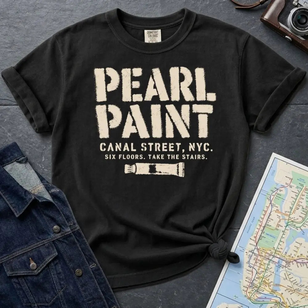 Black Comfort Colors t-shirt with Pearl Paint in large distressed stencil lettering, Canal Street NYC and Six Floors Take The Stairs below, with a paint tube graphic, displayed on a flat surface with a denim jacket and subway map in the background.