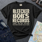 Black Comfort Colors t-shirt with Bleecker Bob's Records in large distressed block lettering, London, New York, Hollywood and Open When Everyone Else Is Closed below, displayed on a flat surface with a denim jacket and subway map in the background.