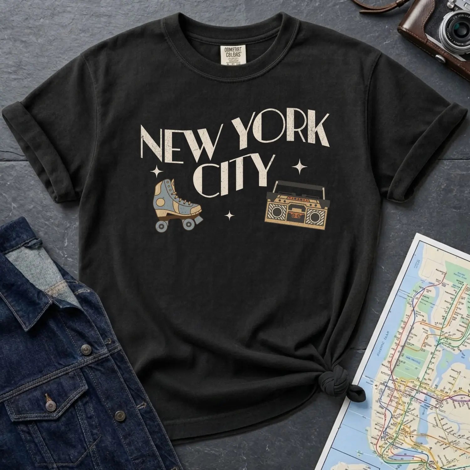 Black Comfort Colors t-shirt with 'New York City' text and a roller skate and boom box on a dark surface with a map and jacket.