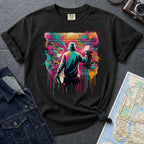 Black Comfort Colors t-shirt with a colorful graphic of a person spray painting a brick wall, placed on a dark surface with a map and denim jacket.