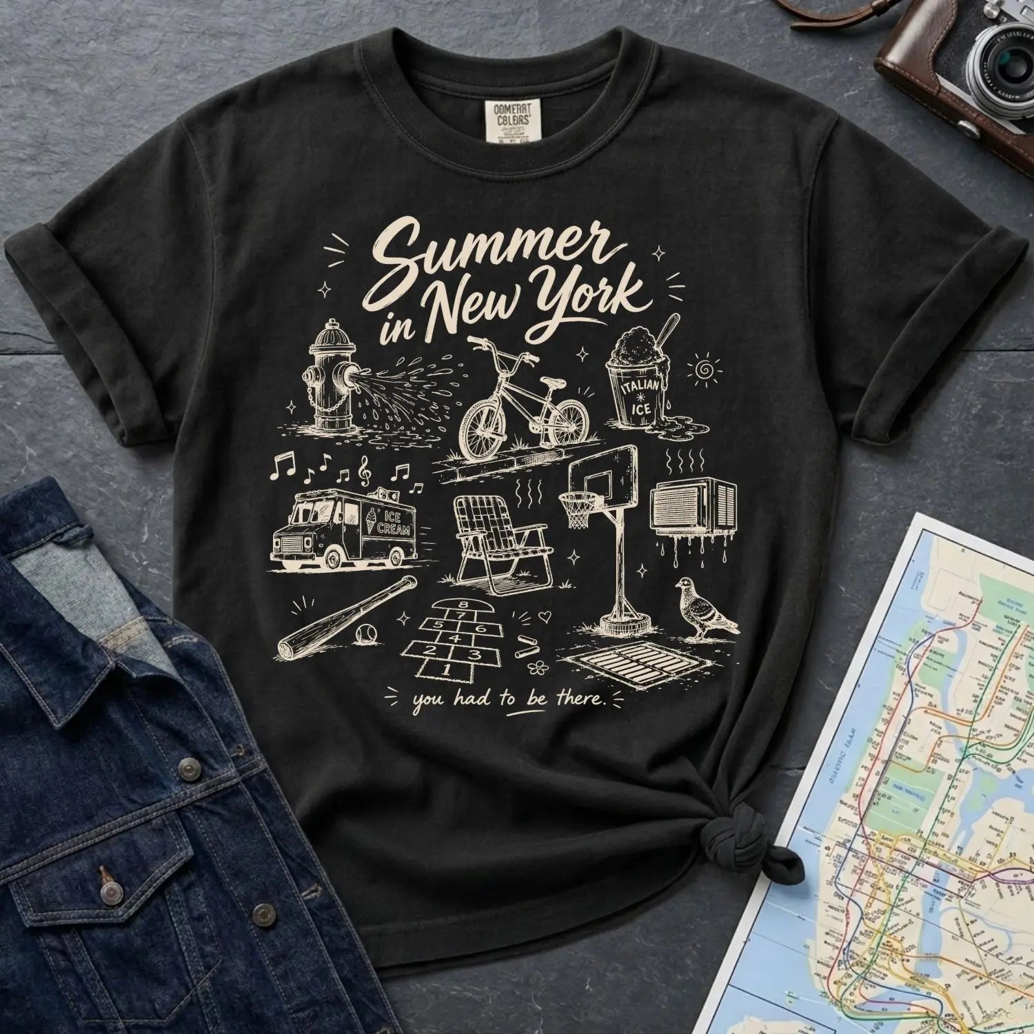 Black Comfort Colors t-shirt with 'Summer in New York' graphic design on a gray surface with denim jacket and map.