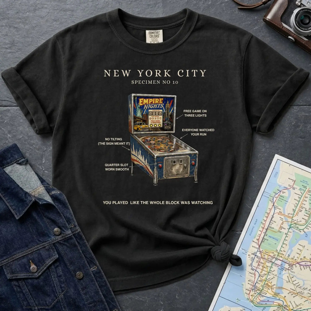 Black Comfort Colors t-shirt with pinball machine graphic and text on a dark surface with a denim jacket and map.