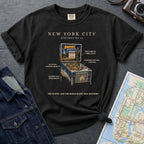 Black Comfort Colors t-shirt with pinball machine graphic and text on a dark surface with a denim jacket and map.