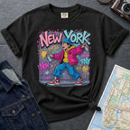 Black Comfort Colors t-shirt featuring a colorful NYC graffiti-style New York graphic with a young street artist holding a spray paint can design on a gray surface with denim jacket and map.