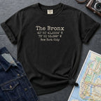 Black Comfort Colors t-shirt with 'The Bronx' coordinates text on a dark surface with a map and jacket.