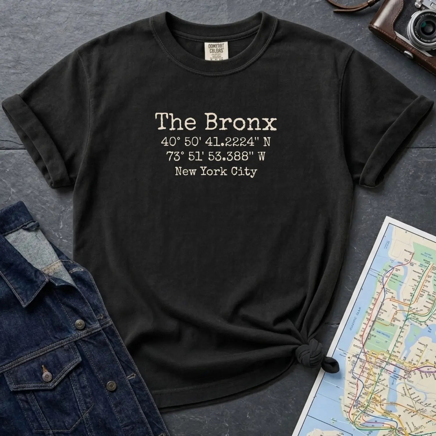 Black Comfort Colors t-shirt with 'The Bronx' coordinates text on a dark surface with a map and jacket.