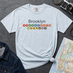 Light blue t-shirt with 'Brooklyn' and subway line numbers on a dark surface with a map and camera.