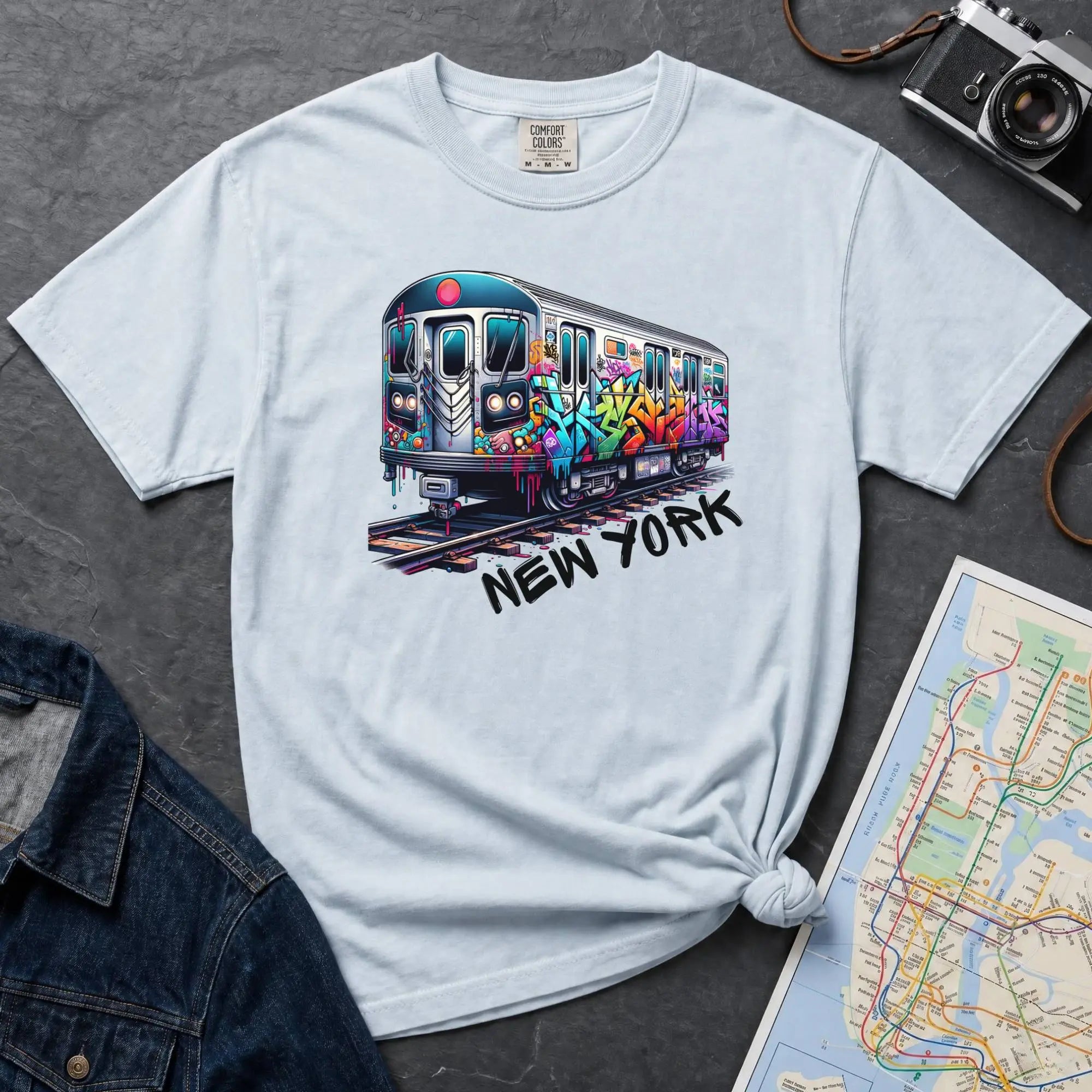 Light blue t-shirt with a colorful train graphic and 'New York' text on a gray surface with a map and denim jacket.