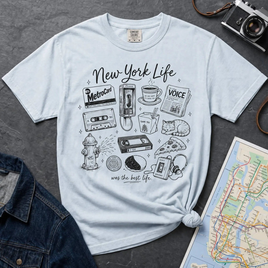 Light blue t-shirt with New York-themed graphics on a gray surface with a map and camera.
