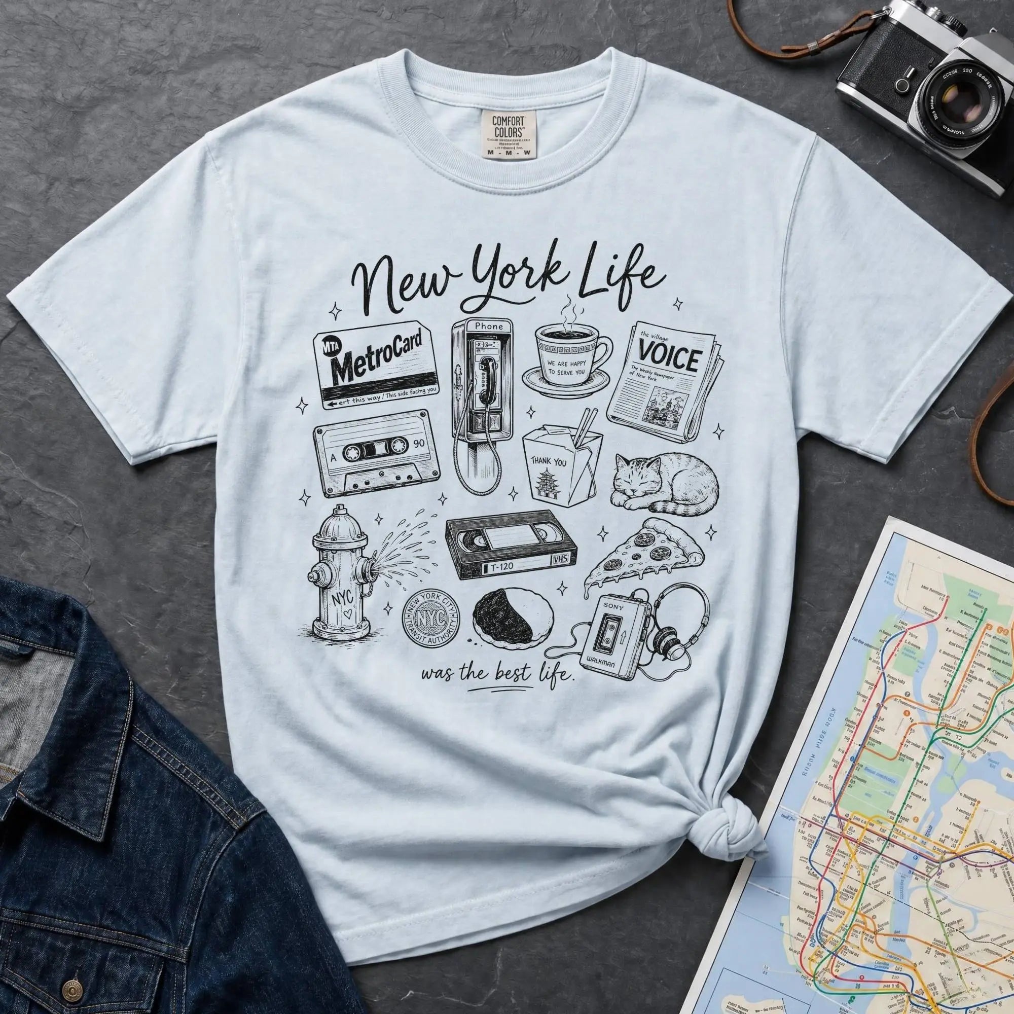 Light blue t-shirt with New York-themed graphics on a gray surface with a map and camera.