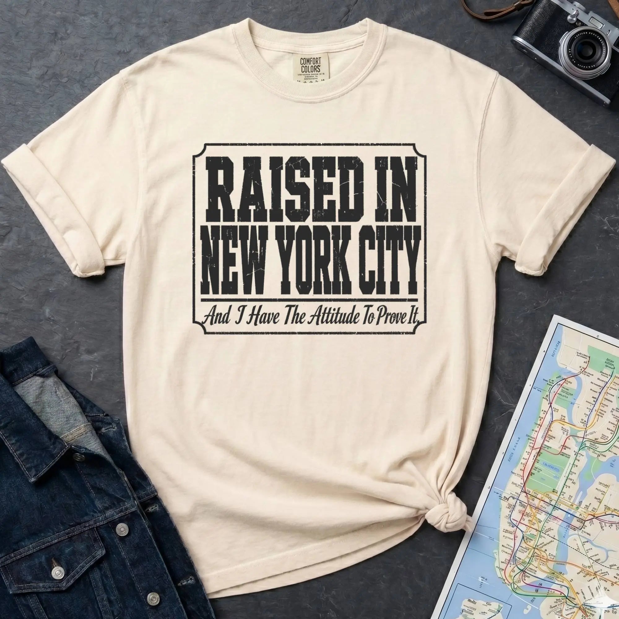 Ivory t-shirt with 'Raised in New York City' text on a dark surface with a jean jacket and a map.