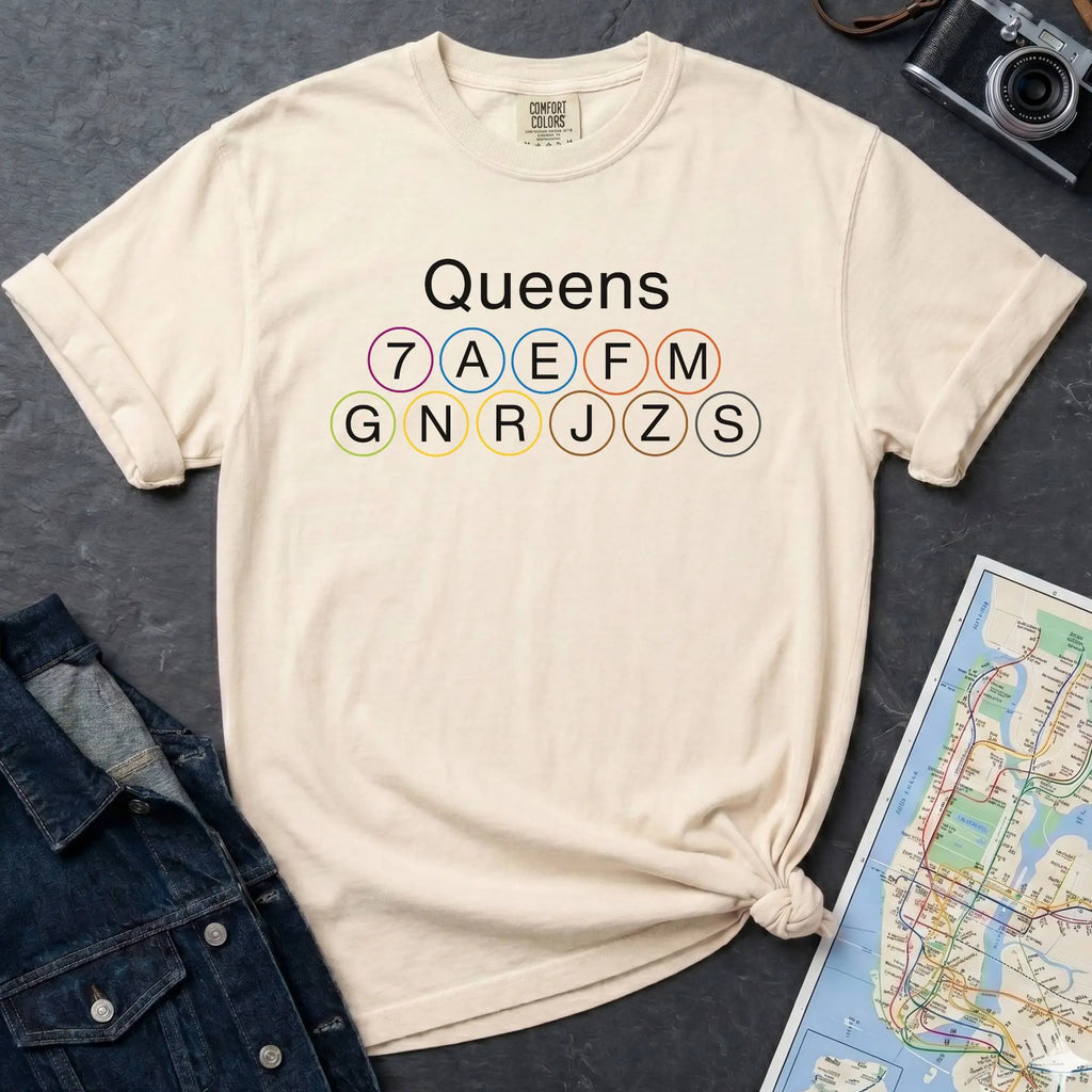 Ivory t-shirt with 'Queens' and subway line numbers on a dark surface with a map and camera.