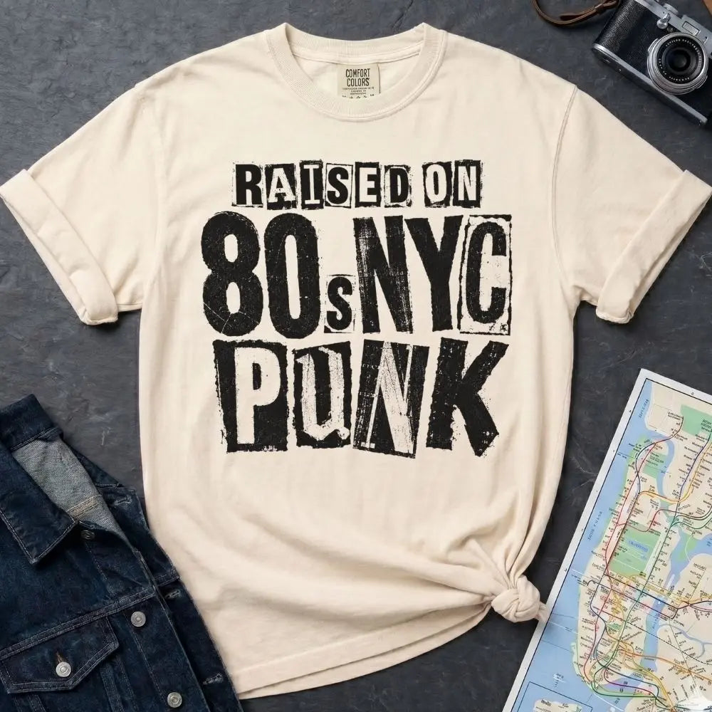 Ivory Comfort Colors t-shirt with bold distressed ransom note style lettering reading "Raised on 80s NYC Punk" in mixed fonts and sizes, black ink on ivory, xerox zine aesthetic.