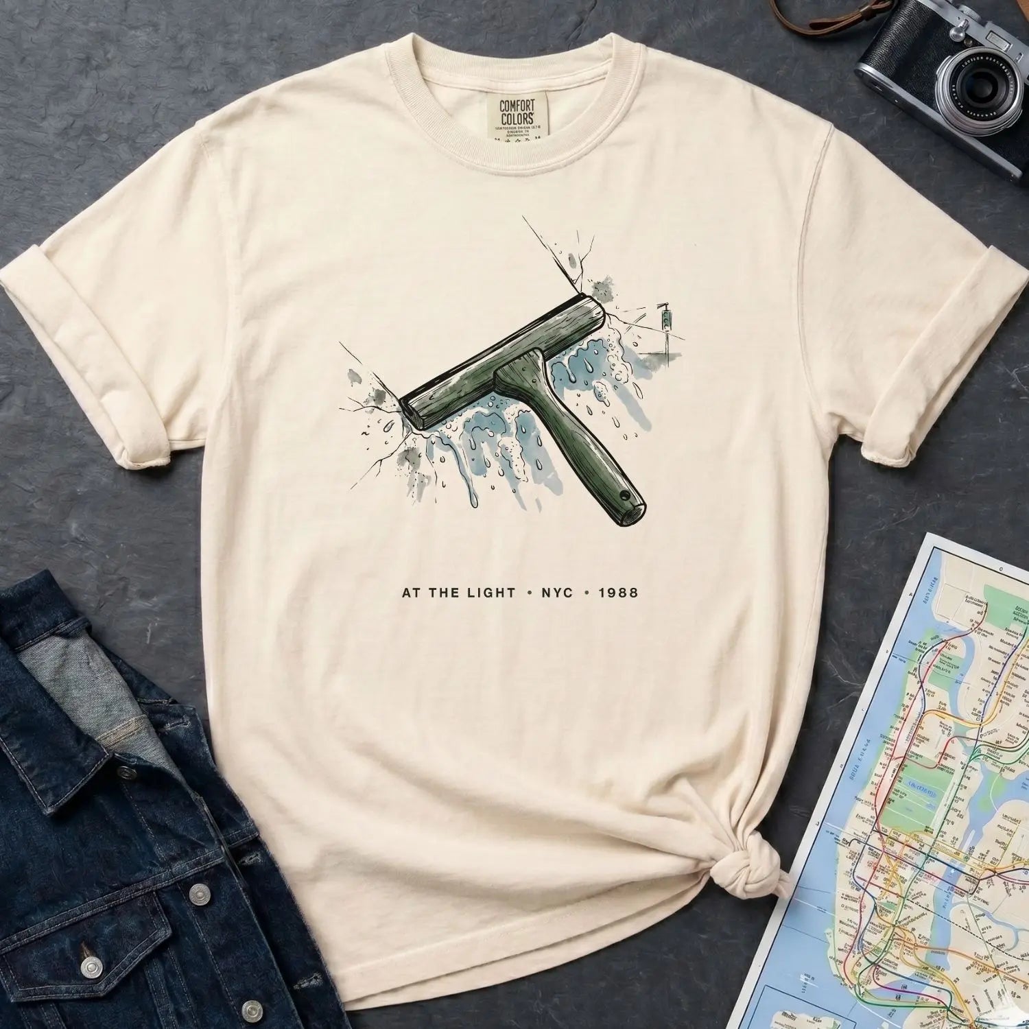 Ivory Comfort Colors t-shirt with graphic design of a squeegee and text, placed on a dark surface with a denim jacket and a map.