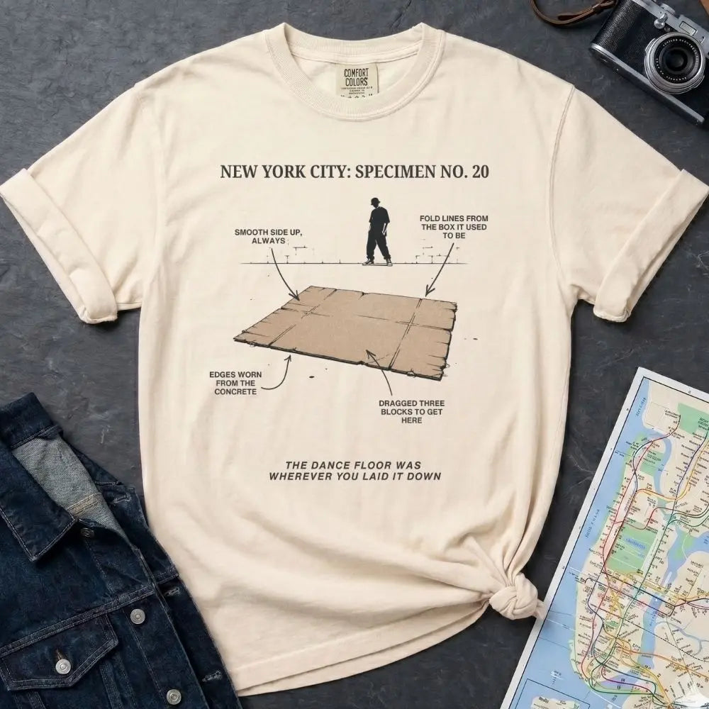 Ivory Comfort Colors T-shirt with NYC breakdance cardboard specimen front print, styled with denim jacket, camera, and subway map.