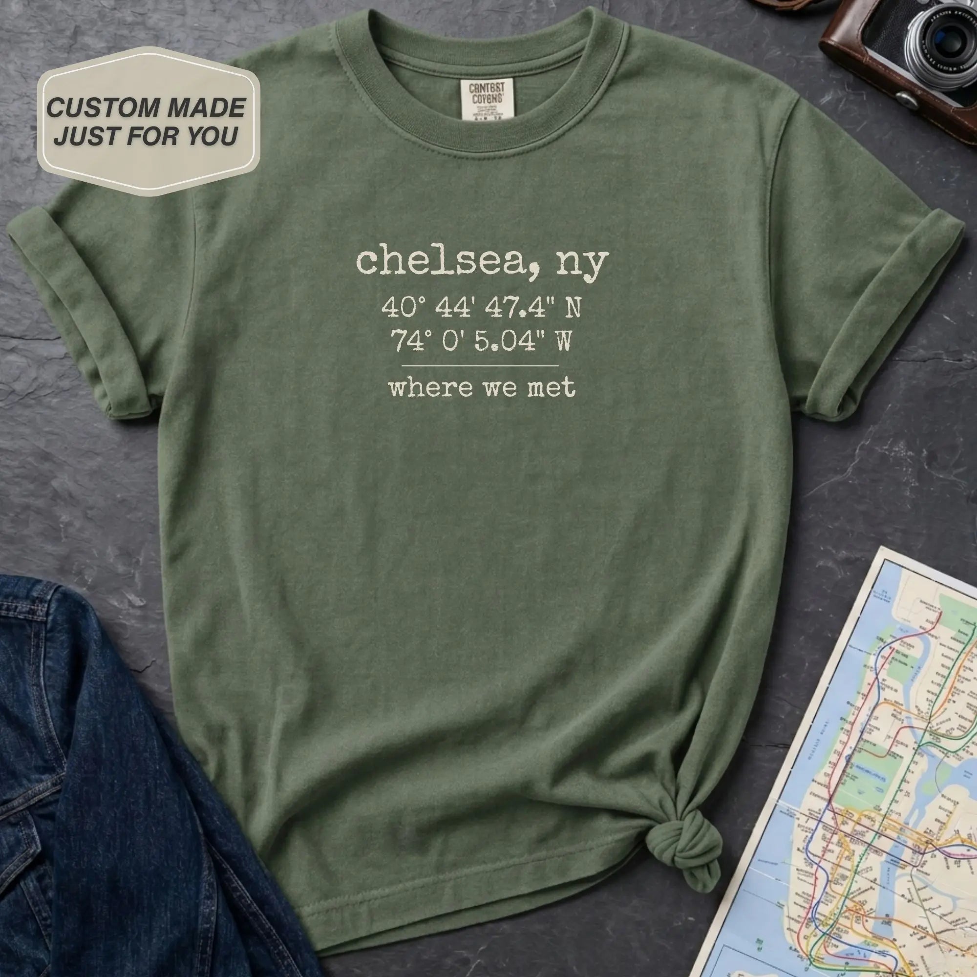 Green t-shirt with coordinates and 'Chelsea, NY' text on a dark surface with a map and jeans.