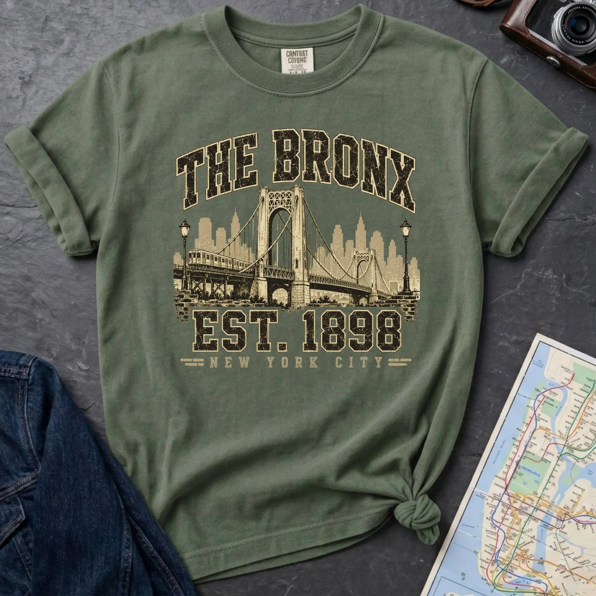 Moss green t-shirt with 'The Bronx Est. 1898' design on a dark surface with a map and a denim jacket.