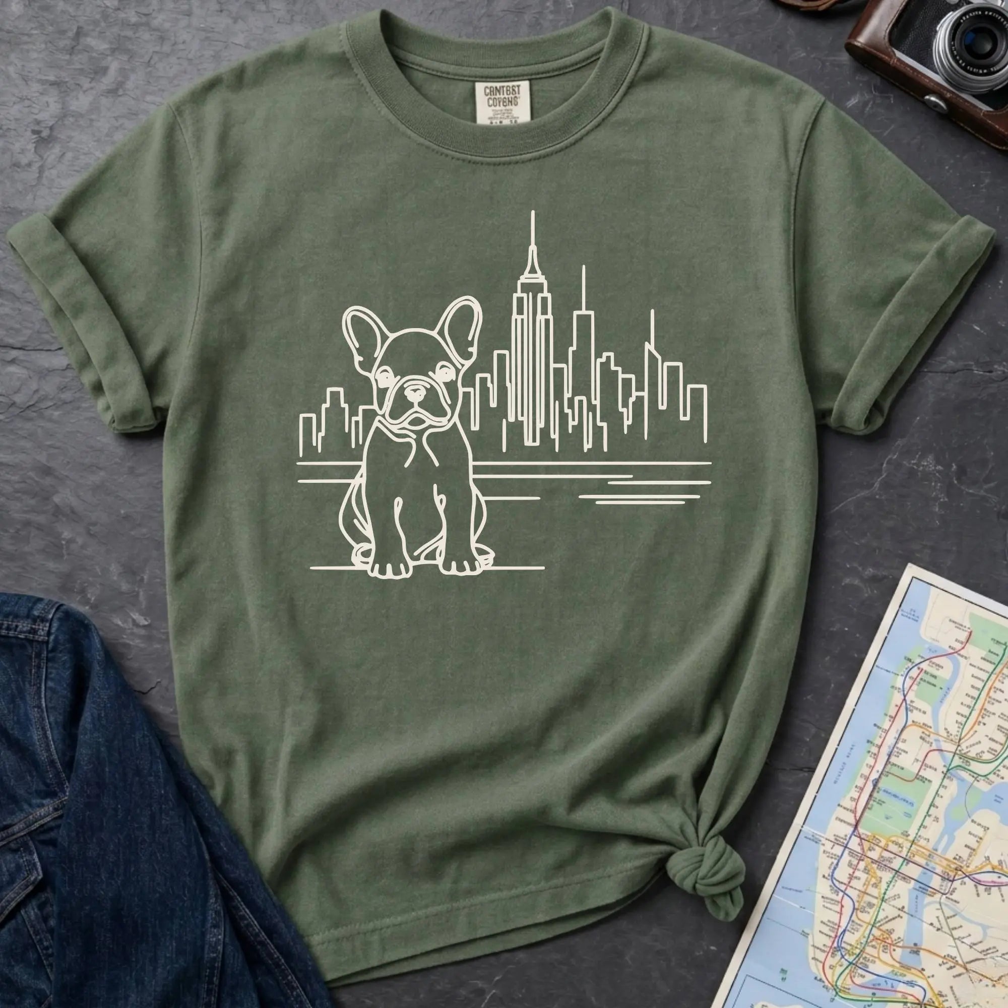Moss green t-shirt with a French Bulldog and cityscape design on a gray surface with a denim jacket and a map.