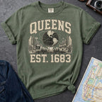 Green t-shirt with 'Queens Est. 1683' graphic design on a dark surface with a denim jacket and a map.