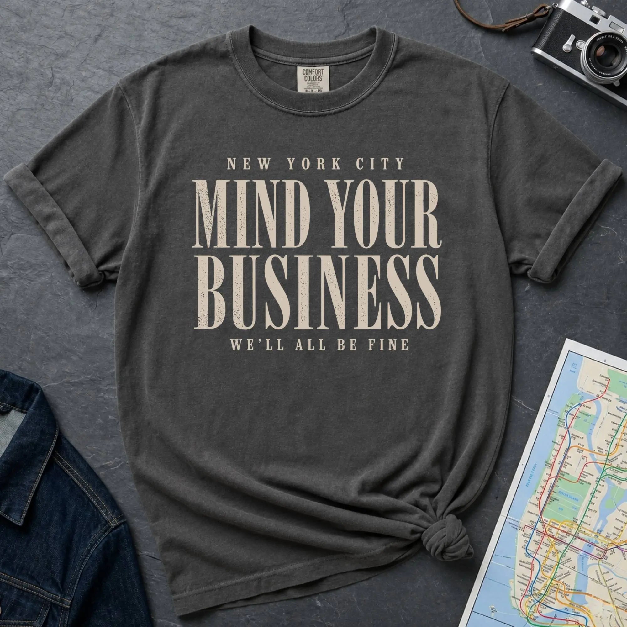 Pepper colored t-shirt with 'New York City Mind Your Business We'll All Be Fine' text on a dark surface with a map and camera.