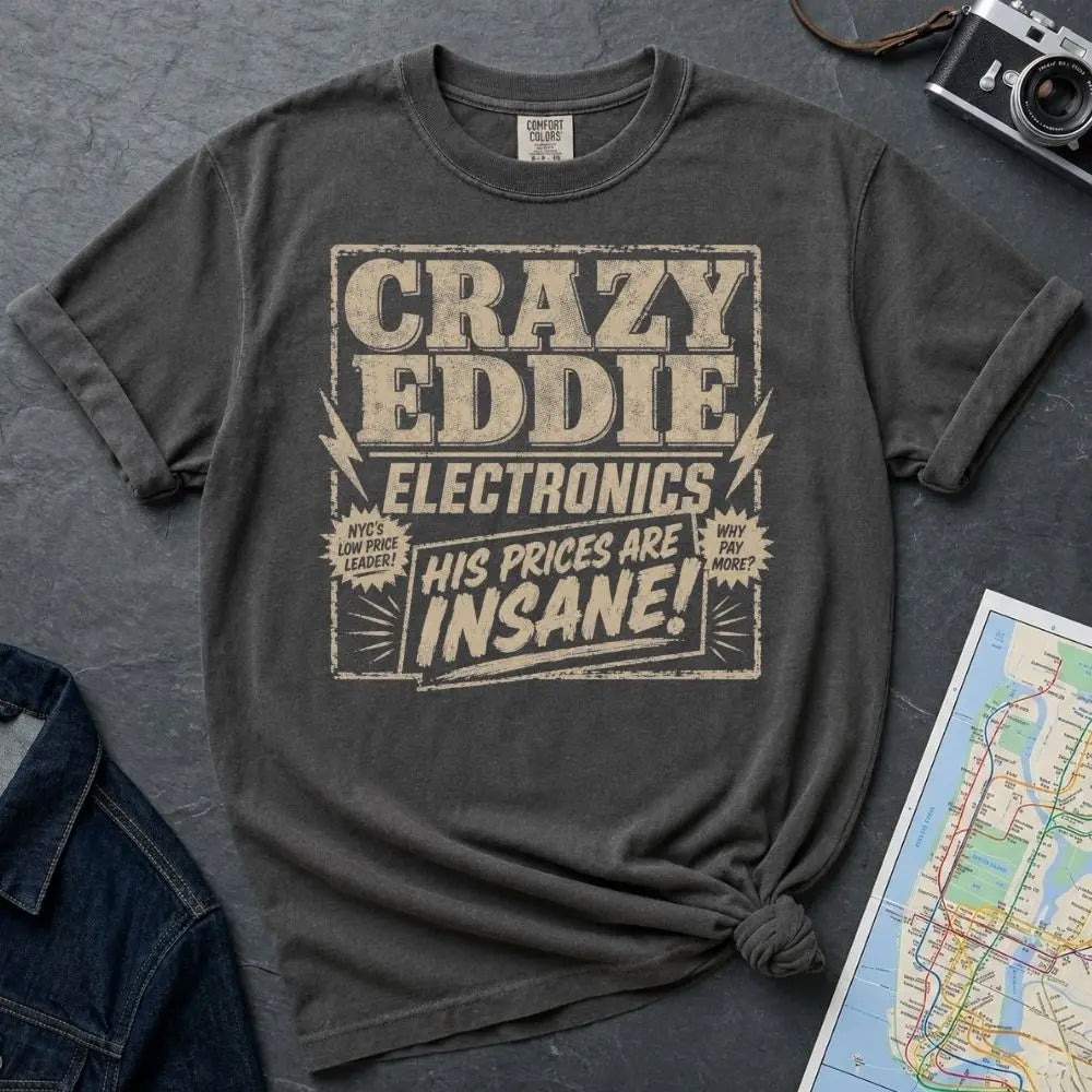 Pepper colored Comfort Colors t-shirt with distressed vintage retail graphic reading Crazy Eddie's Electronics in bold blocky lettering with lightning bolts and the tagline His Prices Are Insane, styled after 1980s NYC consumer electronics store signage.