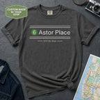 Dark gray t-shirt with '6 Astor Place Still My Stop' text on a textured surface with a map and camera.